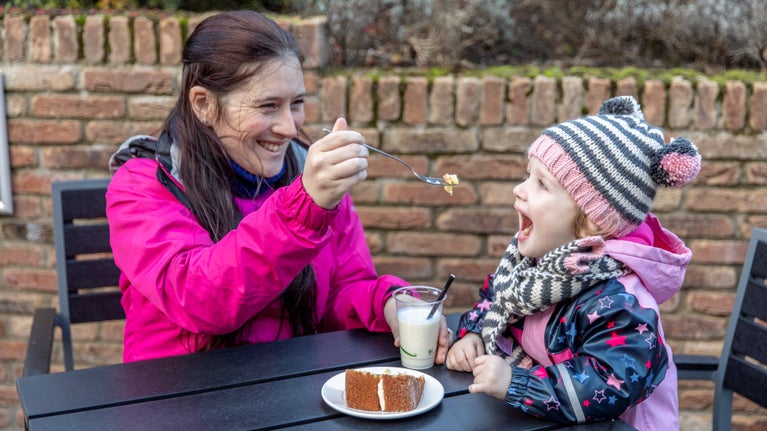 A woman feeding a young girl in a hat cake sat at a table.